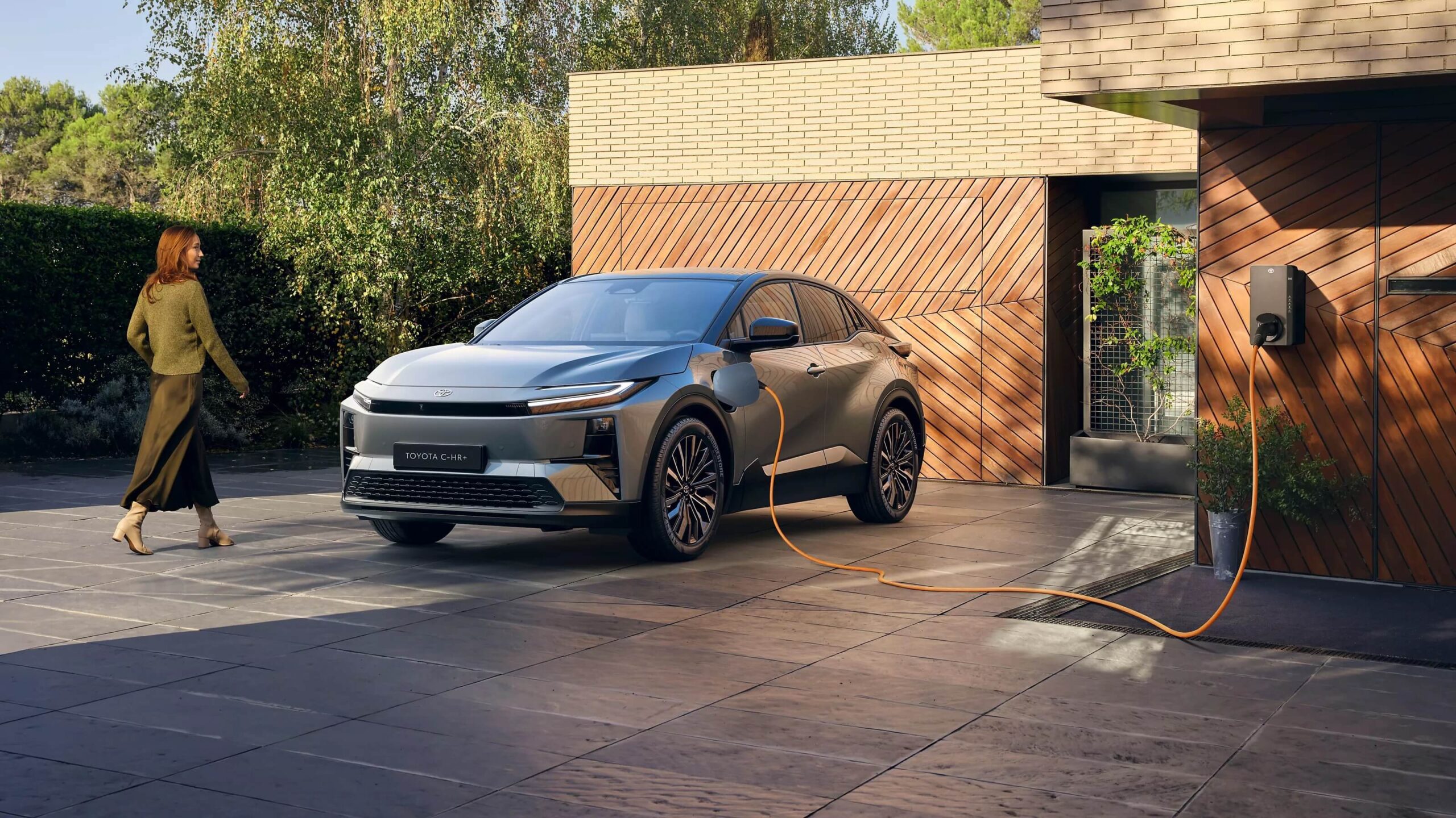 Szary crossover plug-in Toyota C-HR ładowany z domowego wallboxa zamontowanego na drewnianej elewacji, kobieta podchodzi do auta – Toyota Walder Zabrze.